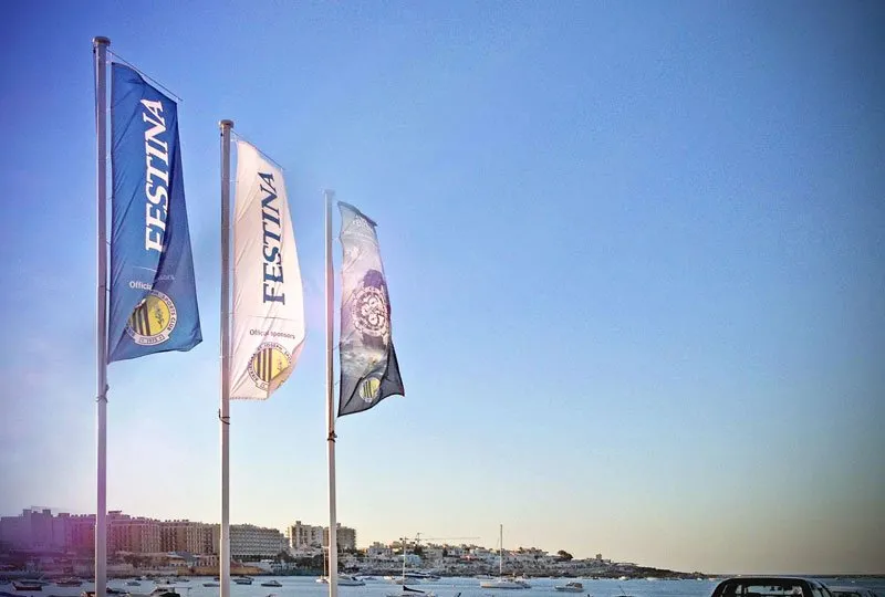 Three printed feather flags flying on poles above a Maltese harbour at dusk