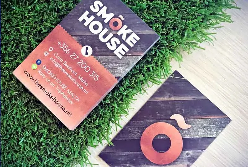 Two printed Smoke House business cards resting on grass and pale wood